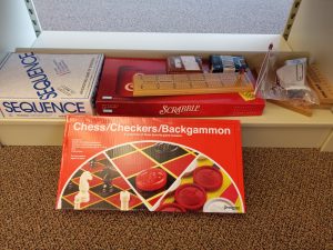 The image displays several board games and a card game positioned on two levels of a shelf. On the top shelf, from left to right, there is a box for the game 